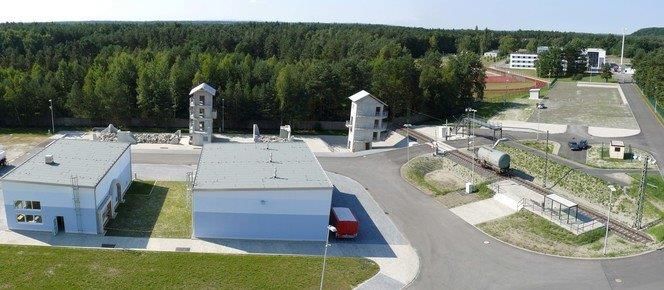 Von oben fotografiert- Zwei weiße Gebäude stehen nahe an asphaltierten Weg, dahinter Wald 